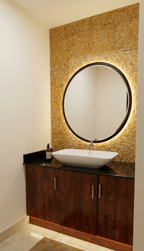 Elegant modern wash area at Riverside Retreat featuring a backlit round mirror on a textured stone wall, a white vessel sink, and a dark wood vanity.